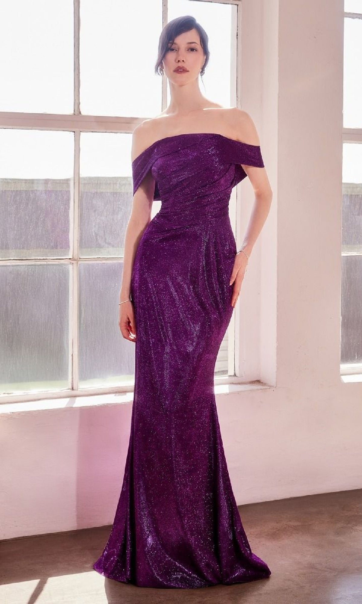 A woman stands indoors by large windows, wearing the Amoris T603—an off-shoulder long glitter prom dress with a sparkling purple mermaid skirt that flows to the floor.